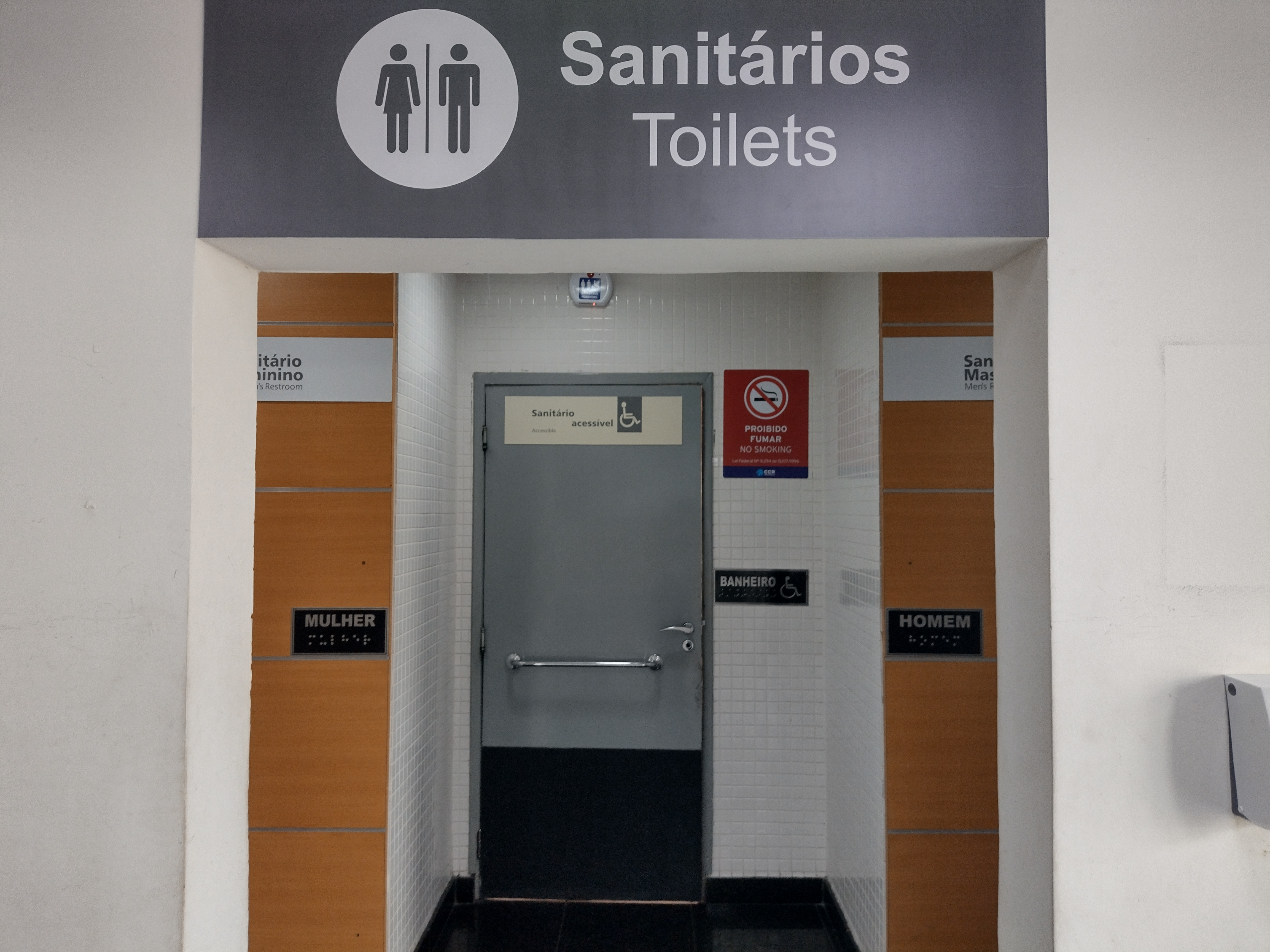 Imagem da entrada de sanitários com sinalização visual e tátil. Ao centro, uma porta com placa “Sanitário acessível” e símbolo de acessibilidade. À esquerda e à direita, entradas para sanitário feminino e masculino com identificação em braille.