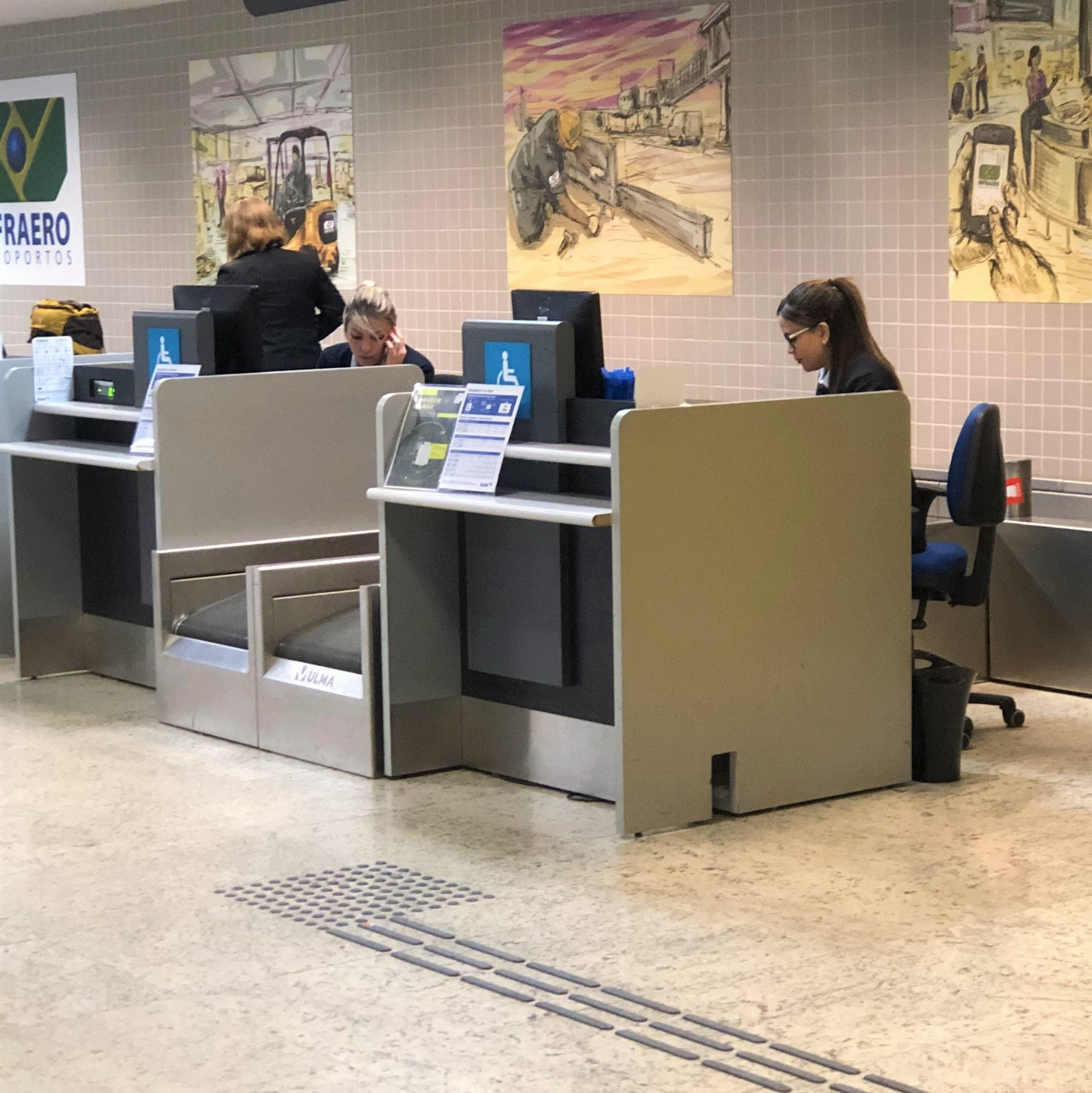 Fotografia de um balcão de atendimento da Infraero com sinalização de acessibilidade. O balcão tem rebaixo frontal para encaixe de cadeiras de rodas. Há profissionais atendendo e cartazes informativos fixados nas divisórias. O piso tátil direcional passa em frente aos balcões, indicando o caminho.