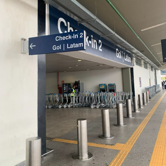 Imagem da entrada do setor de check-in 2 do aeroporto, com letreiro azul suspenso indicando as companhias Gol e Latam. À frente da entrada, há rota tátil em piso amarelo com linha direcional e alerta, além de barreiras metálicas organizando o fluxo. A área interna está equipada com carrinhos de bagagem e máquinas de autoatendimento.