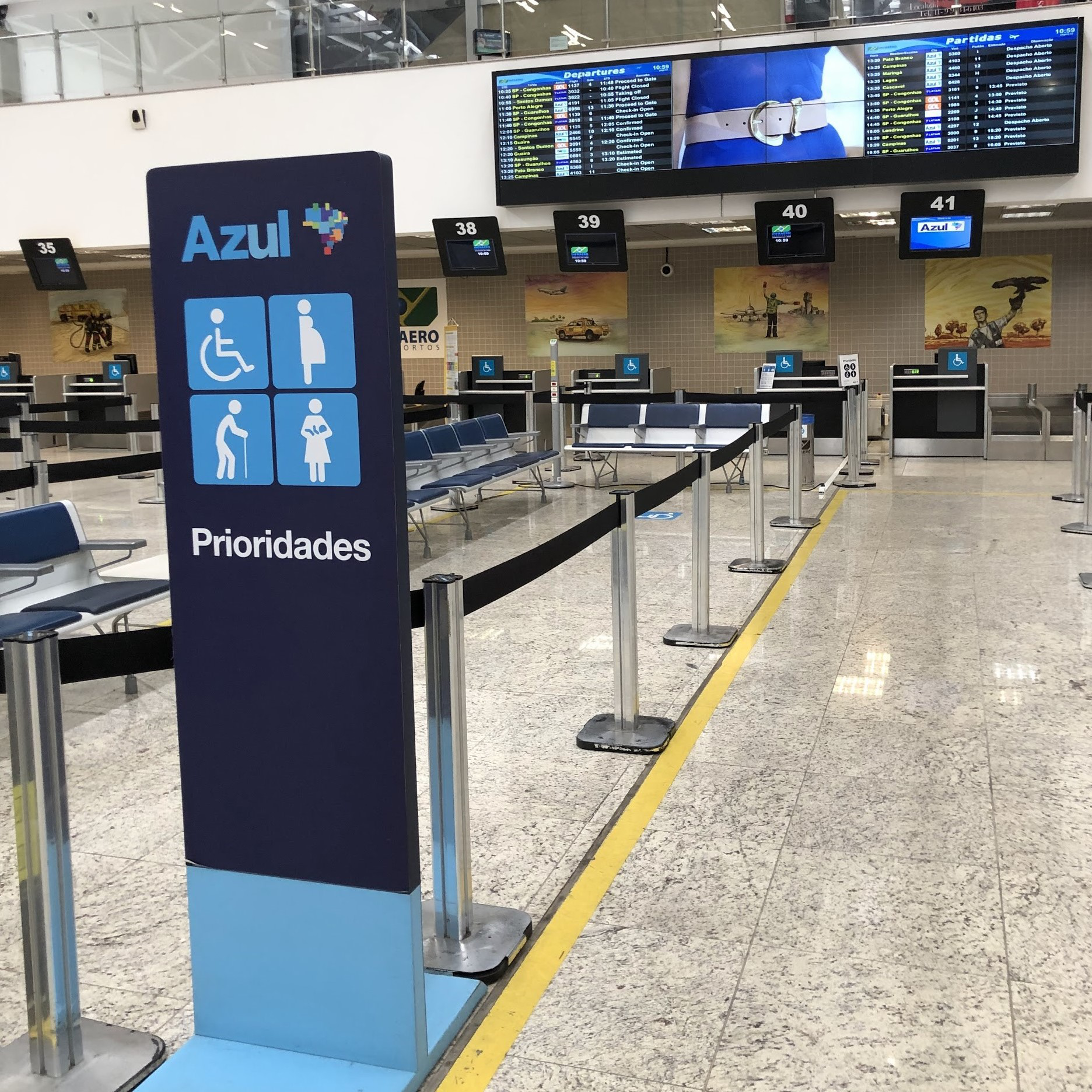 Imagem da área interna do aeroporto com placa vertical destacando o atendimento prioritário da companhia Azul. A placa possui ícones para pessoas com deficiência, gestantes, pessoas idosas e com criança de colo. Ao fundo, estão os balcões de check-in com sinalização de acessibilidade e painéis de voos suspensos no teto.