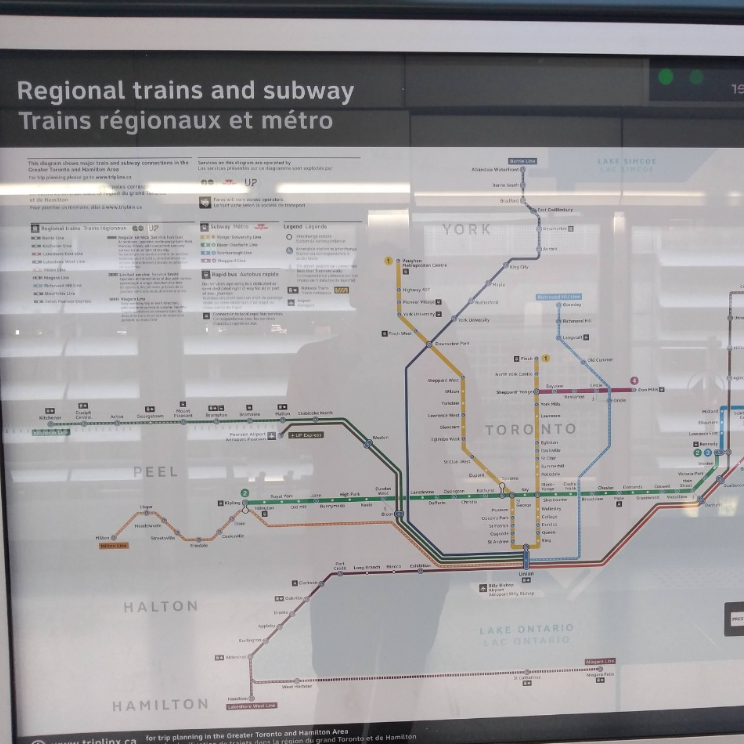 Painel gráfico com fundo branco apresentando o diagrama do sistema de transporte público da região de Toronto, no Canadá. Estão representadas linhas coloridas que indicam trajetos de trens e metrô, com legendas e paradas ao longo dos itinerários. Acima do mapa, os textos estão bilíngues, em inglês e francês.