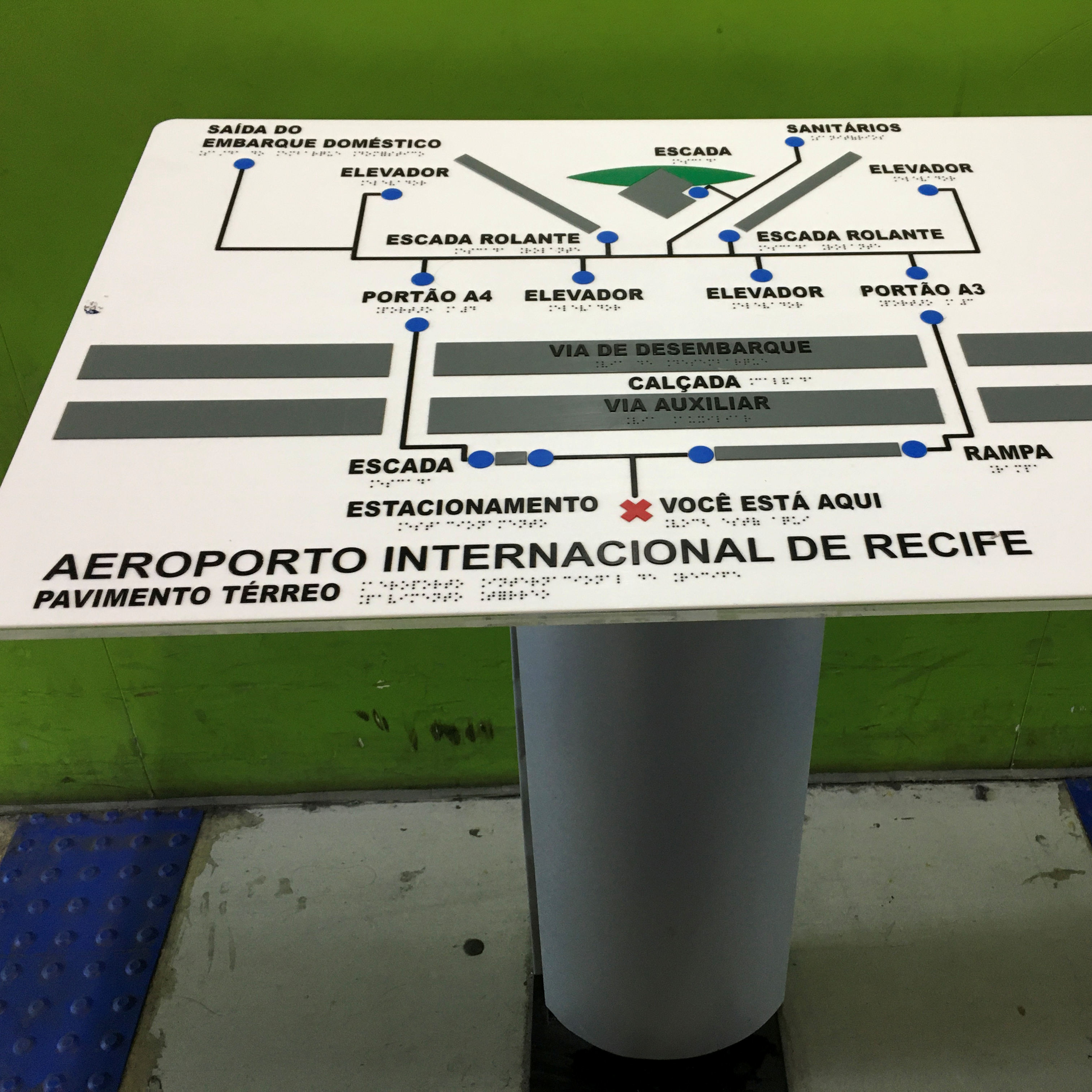 Imagem de um mapa tátil do Aeroporto Internacional de Recife. O mapa está posicionado sobre um pedestal e apresenta rotas, elevadores, escadas e rampas com sinalização em Braille e relevo. Há uma indicação clara de "VOCÊ ESTÁ AQUI" com um X vermelho em alto relevo. O fundo é verde e o piso possui sinalização tátil direcional.