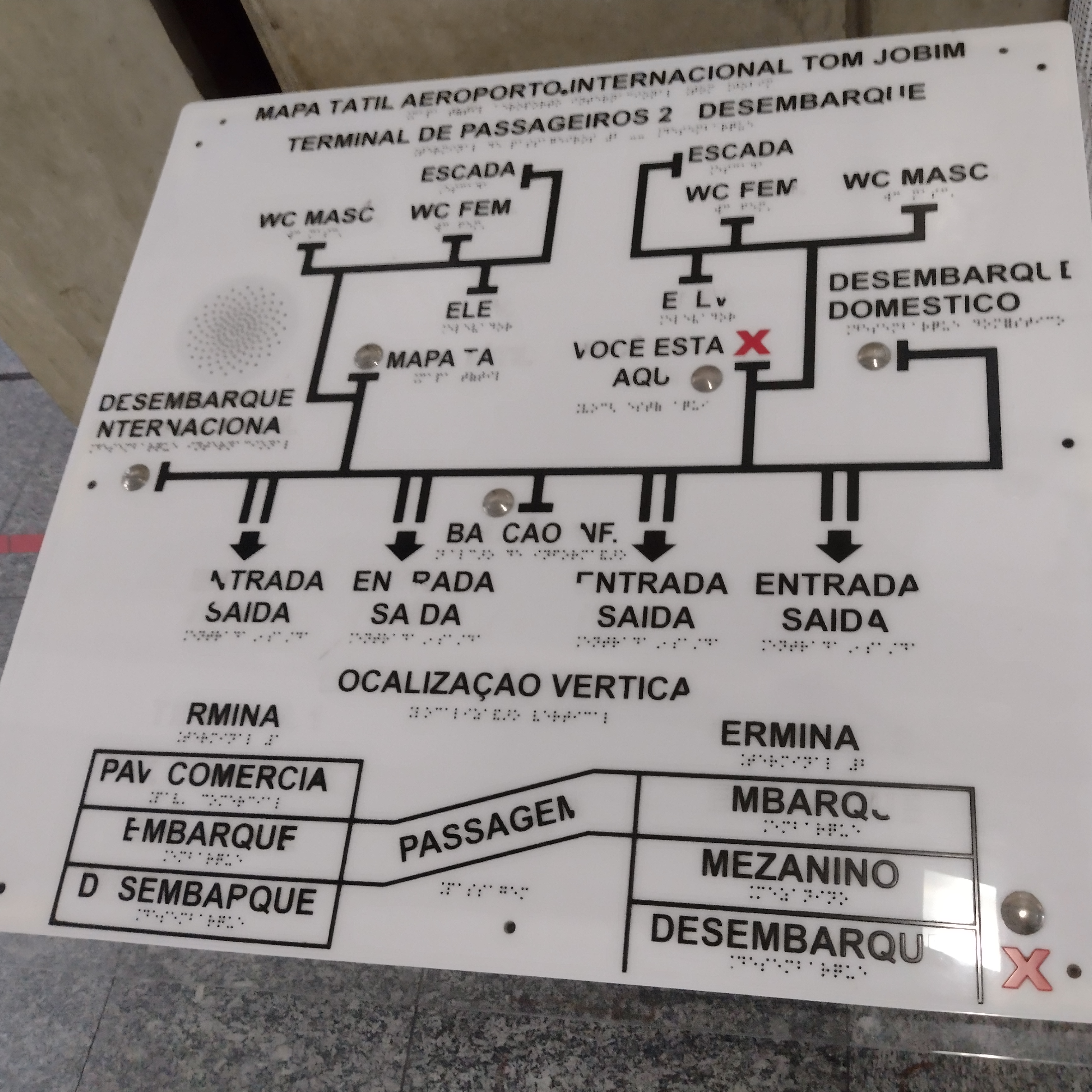 Mapa tátil do Aeroporto Internacional Tom Jobim (Galeão – RJ). O painel exibe planta baixa do terminal com caminhos, elevadores, sanitários e áreas de embarque/desembarque. Todos os textos estão acompanhados por escrita em Braille. Uma marca vermelha em forma de X indica a posição atual do usuário. O material é branco com linhas pretas em relevo.