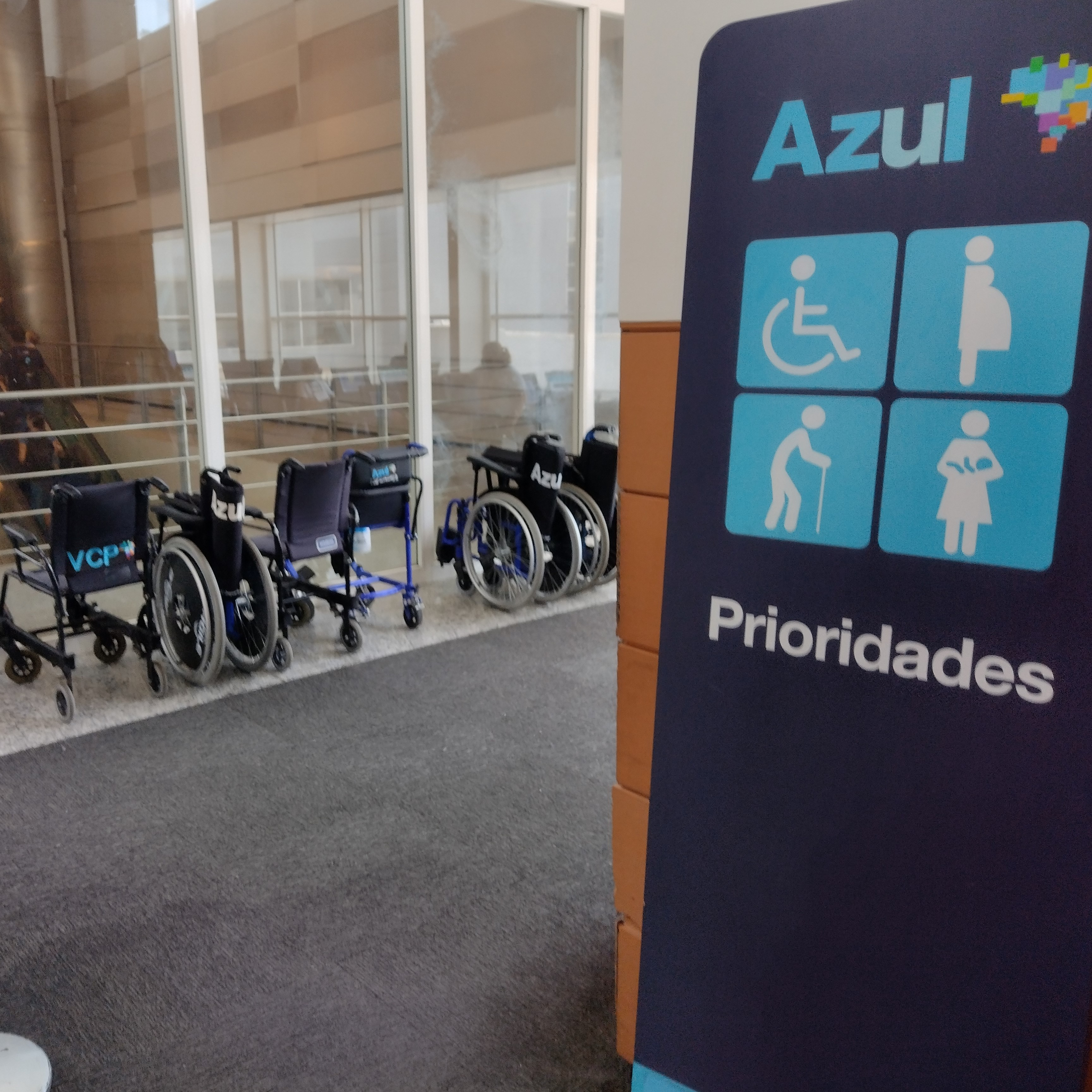 Placa de sinalização vertical da companhia aérea Azul indicando "Prioridades". A placa exibe quatro pictogramas azuis e brancos: pessoa em cadeira de rodas, pessoa gestante, pessoa idosa com bengala e pessoa com criança de colo. Ao lado da placa, diversas cadeiras de rodas estão enfileiradas próximo a uma parede de vidro.