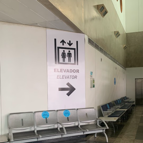 Placa de sinalização vertical em uma parede branca indicando a direção de um elevador. A placa exibe o pictograma de elevador com setas para cima e para baixo, as palavras "ELEVADOR" e "ELEVATOR", e uma seta grande apontando para a direita. Abaixo da placa, há uma fileira de assentos metálicos com adesivos azuis indicando distanciamento.