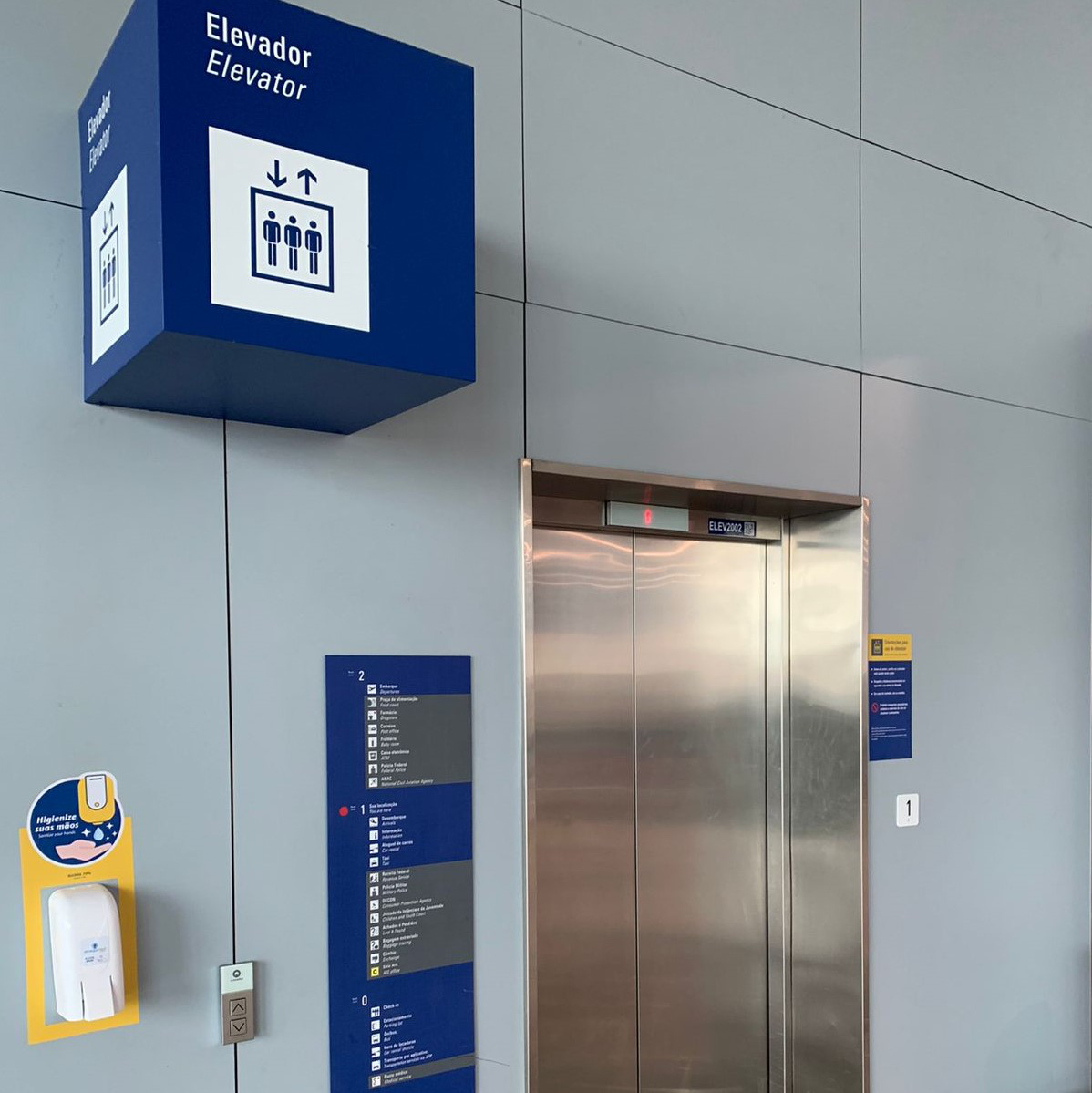 Sinalização indicativa para elevador em um ambiente interno com paredes cinzas. Uma placa azul em formato de cubo, suspensa, exibe o pictograma de elevador e as palavras "Elevador" e "Elevator". Ao lado, as portas metálicas de um elevador. Próximo ao elevador, há um totem com informações e botões de chamada. Um dispenser de álcool em gel está fixado na parede.