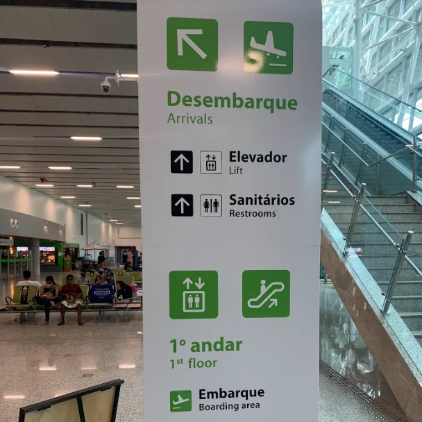 Painel de sinalização vertical em um aeroporto, indicando a localização de diversos serviços. Setas e pictogramas direcionam para "Desembarque/Arrivals", "Elevador/Lift", "Sanitários/Restrooms", "1º andar/1st floor" e "Embarque/Boarding area". Ao fundo e à direita, uma escada rolante é visível.