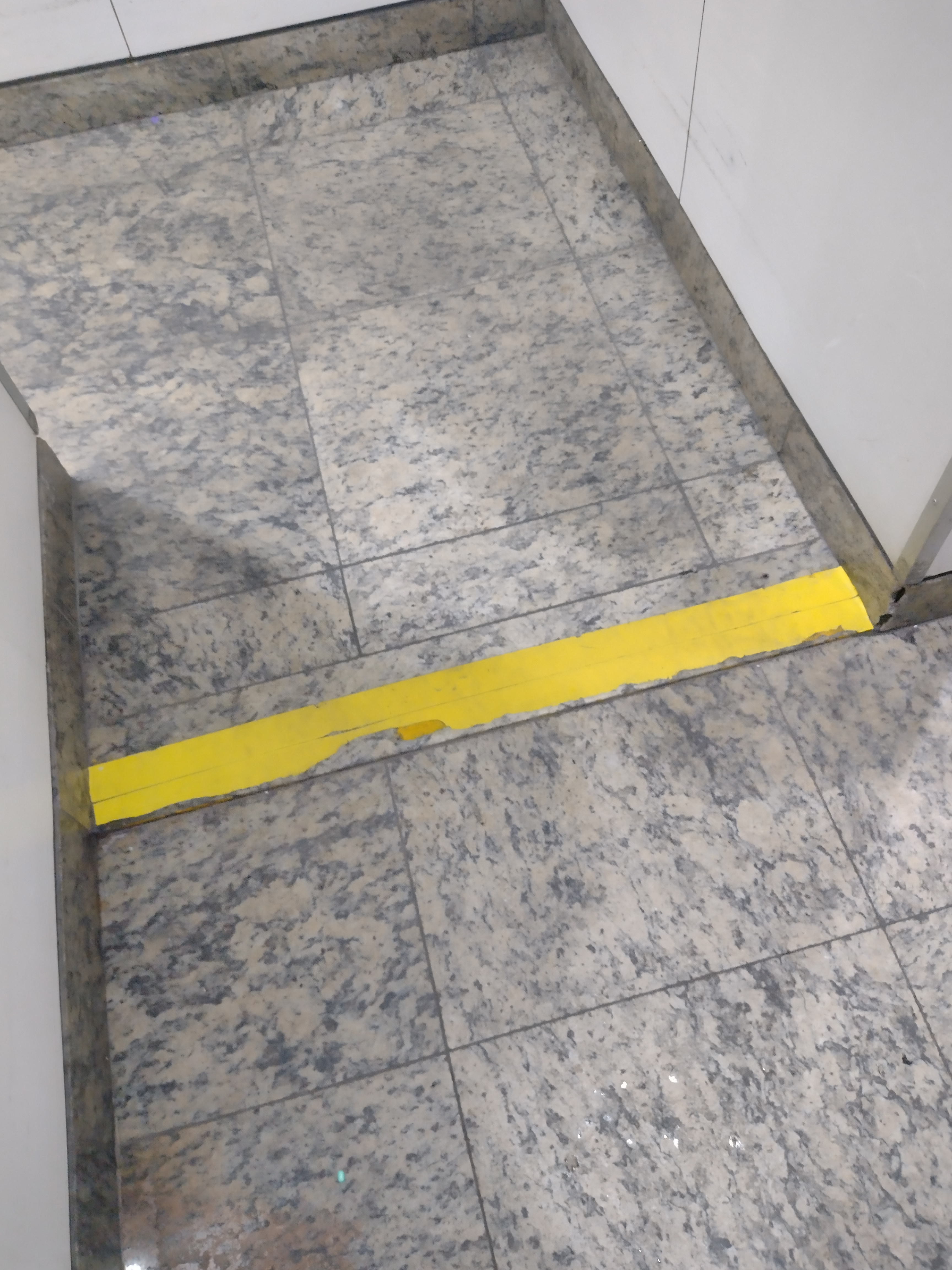 Piso de granito com faixa adesiva amarela em contraste, aplicada na borda de um pequeno desnível dentro de um elevador, alertando visualmente sobre a transição entre níveis.