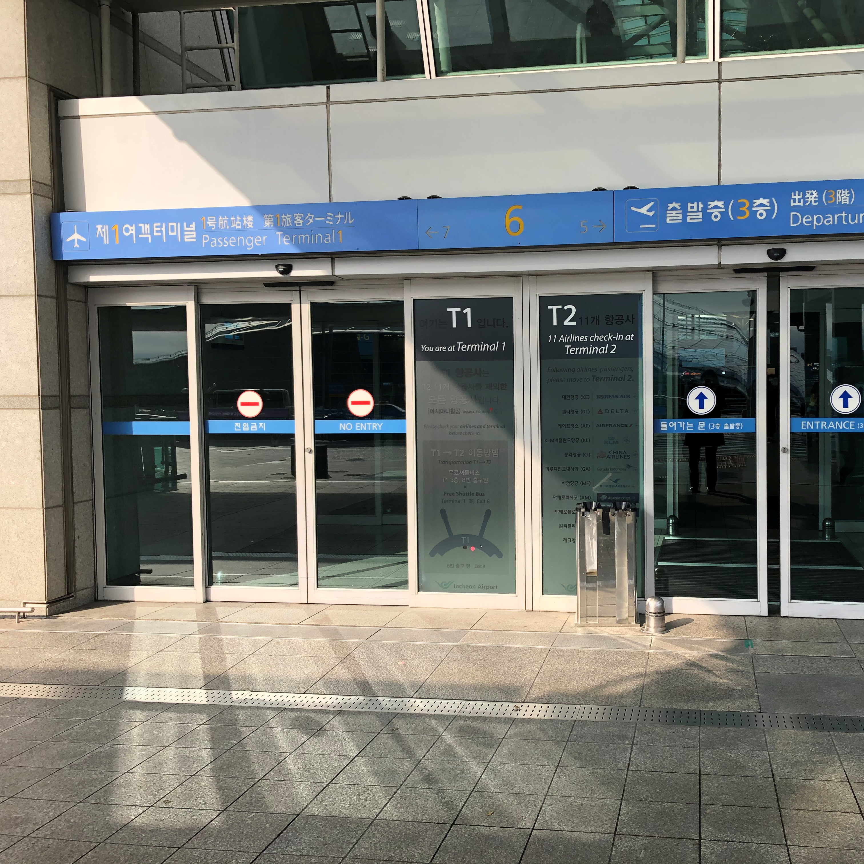 Portas de vidro automáticas de entrada de um terminal de passageiros (Passenger Terminal 1 e 2). As portas possuem faixas horizontais azuis com a indicação "NO ENTRY" e círculos vermelhos de proibição. Há também adesivos com informações e o logo "T1" e "T2". Acima das portas, uma longa placa azul contém informações em coreano e inglês.