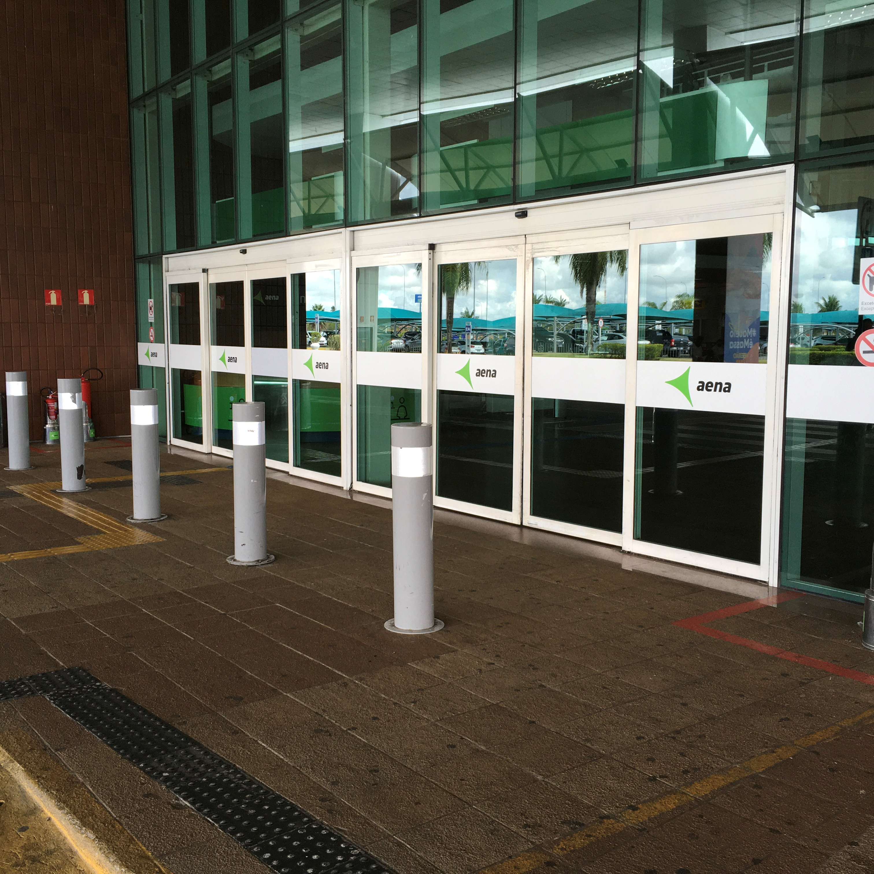 Conjunto de portas de vidro automáticas na entrada de um edifício com fachada de vidro e tijolos aparentes. As portas possuem faixas brancas horizontais com o logo da "aena" para sinalização visual. Balizadores cinzas estão dispostos em frente às portas e piso tátil de alerta demarca a área de circulação.