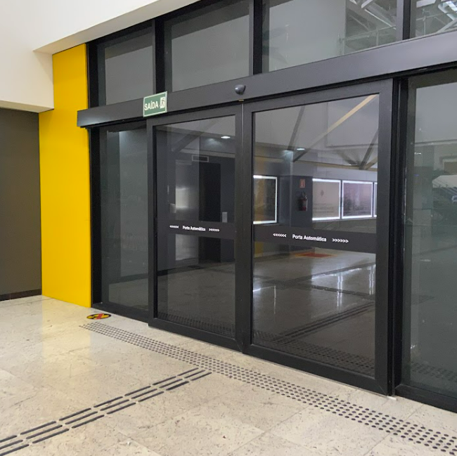 Portas de vidro automáticas com esquadrias pretas em um ambiente interno. As portas possuem faixas brancas horizontais com a inscrição "Porta Automática >>>>>" para sinalização. Uma placa verde com a indicação "SAÍDA" está visível acima das portas. No piso claro, há sinalização tátil de alerta e direcional. Uma parede lateral é pintada de amarelo.