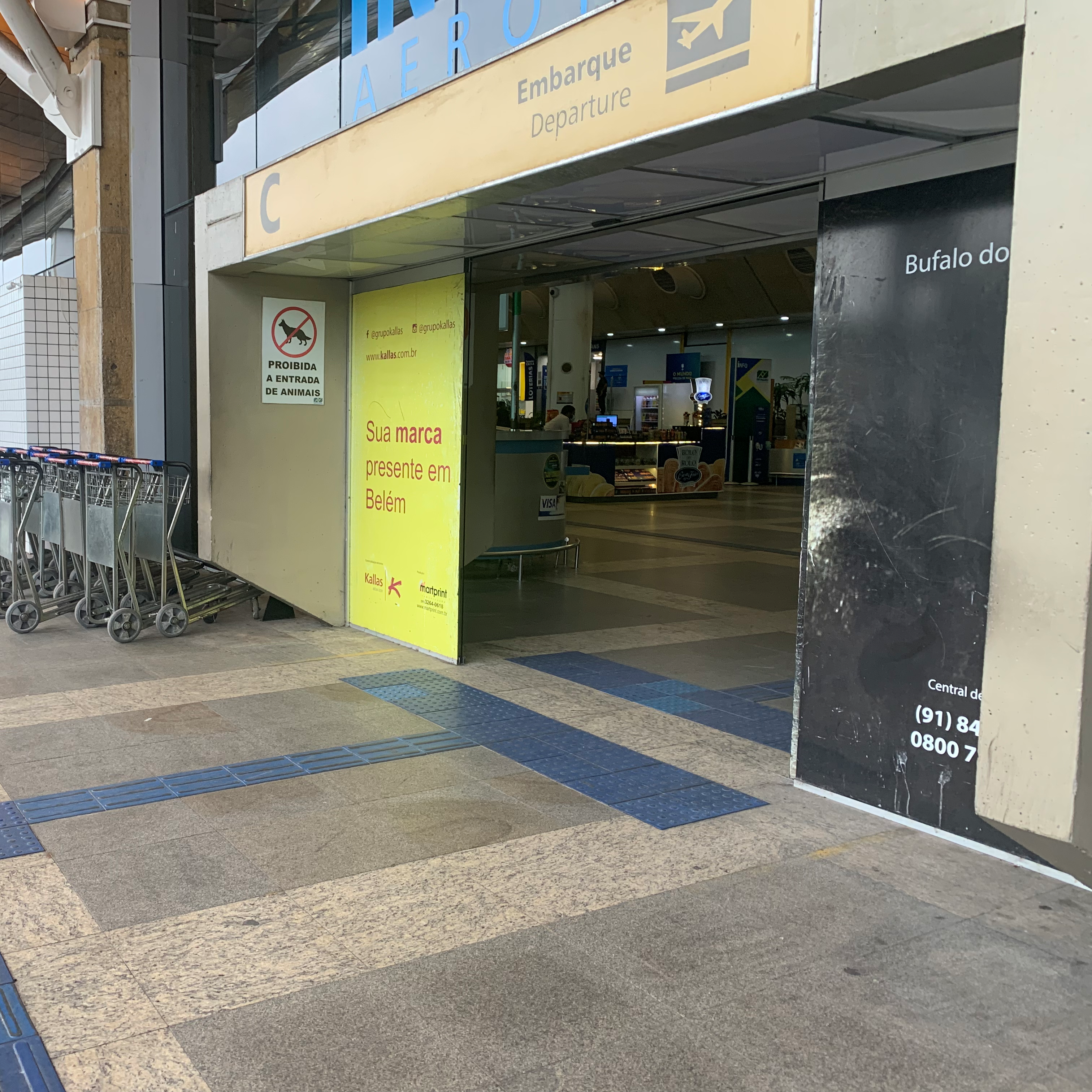 Entrada de aeroporto com piso tátil direcional e de alerta em alto-relevo: a sinalização tátil conduz até a porta automática de entrada, passando por entre dois bloqueios cilíndricos metálicos.