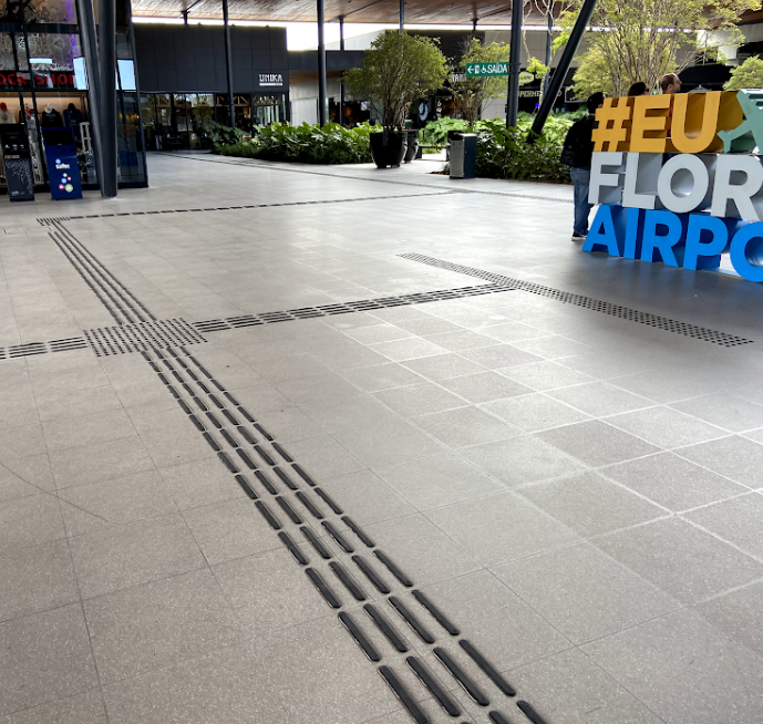 Área em frente ao elevador com piso tátil direcional e de alerta: o piso com linhas em relevo guia até a porta do elevador, e o piso com bolinhas em relevo indica a zona de parada, promovendo orientação precisa para embarque seguro.