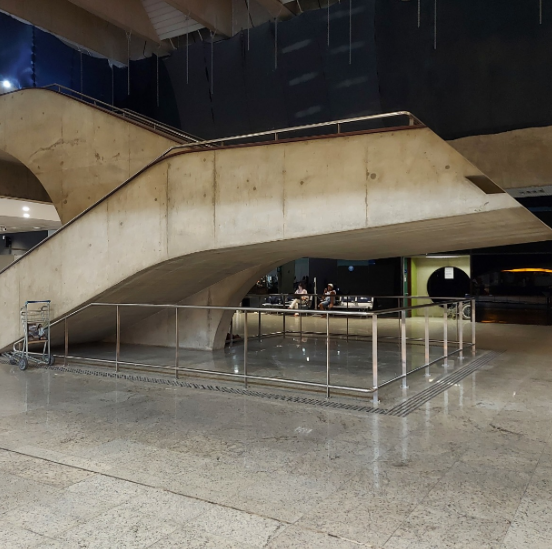 Fotografia de escada com barras de proteção metálicas sob o vão: escada de concreto em área interna de aeroporto com guarda-corpos metálicos delimitando o espaço sob a escada. Ao redor da estrutura, há piso tátil de alerta, indicando o obstáculo suspenso.