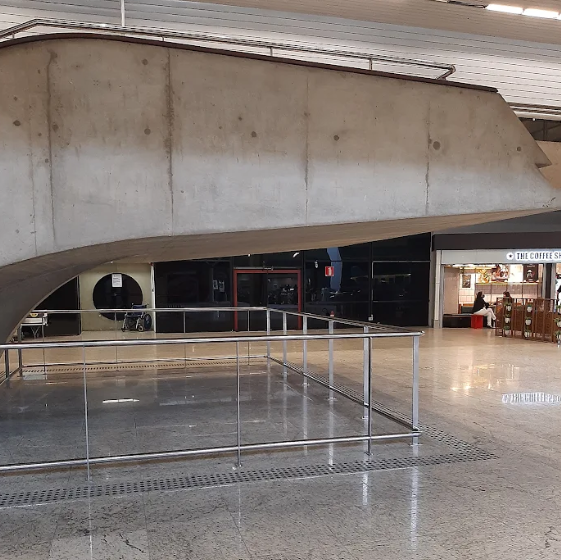 Escada suspensa com proteção metálica perimetral: vista lateral de uma escada em ambiente aeroportuário, onde há uma estrutura metálica cercando a parte inferior da escada, acompanhada de piso tátil de alerta em toda a volta da proteção.