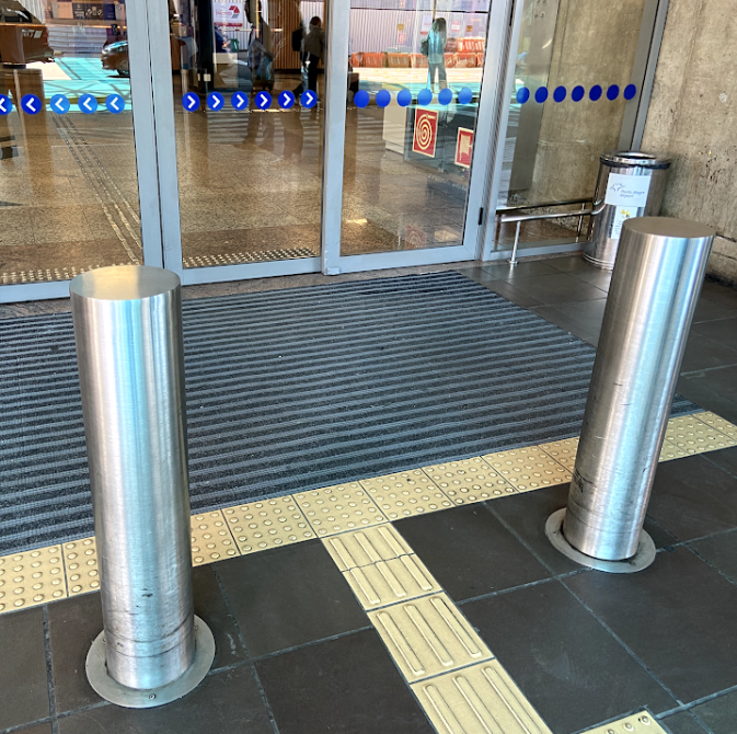 Entrada de porta automática com piso tátil de alerta em frente e piso direcional conduzindo até a porta. Há dois balizadores metálicos que delimitam a passagem, e o caminho tátil está devidamente alinhado para orientar pessoas com deficiência visual.
