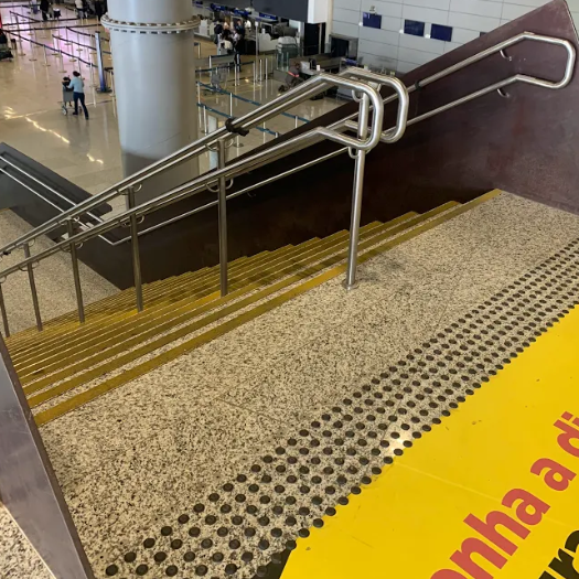 Piso tátil de alerta instalado no topo de uma escada fixa em aeroporto. As bolinhas em relevo indicam a presença do desnível à frente, alertando pessoas com deficiência visual antes da descida. A escada também conta com corrimãos em altura dupla.