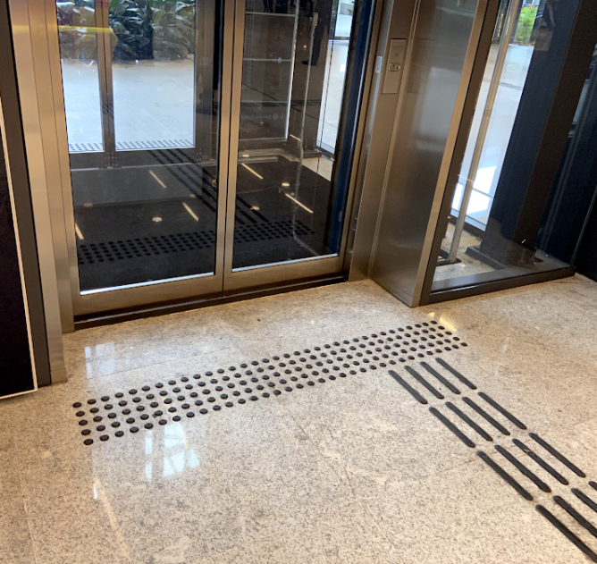 Área em frente ao elevador com piso tátil de alerta (bolinhas em relevo) instalado perpendicularmente à porta e piso tátil direcional (barras em relevo) conduzindo até ele. O padrão de instalação está adequado à norma de acessibilidade.