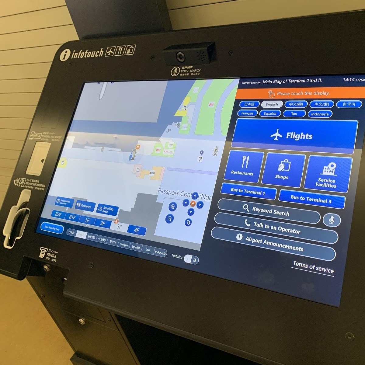 Totem eletrônico de informação acessível, com tela sensível ao toque exibindo um mapa de terminal de aeroporto e opções de navegação por voz, fala com operador e serviços do aeroporto, além de idiomas variados. Ao lado da tela há um fone acoplado para acessibilidade auditiva.