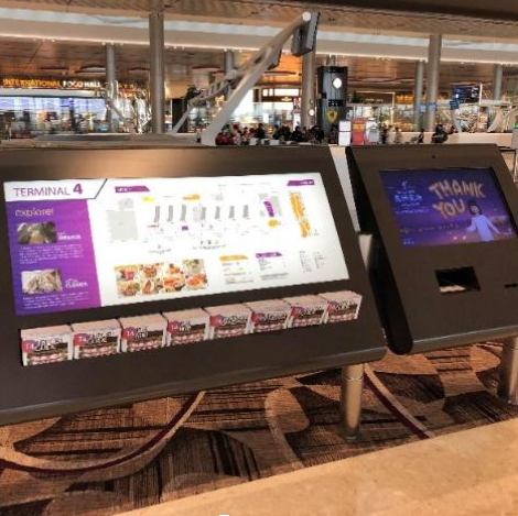 Painel digital interativo do Terminal 4 de um aeroporto internacional, com telas horizontais em posição acessível. Uma das telas exibe informações sobre estabelecimentos, com interface gráfica colorida e acessível ao toque. Ao lado, há outra tela com mensagem de agradecimento.