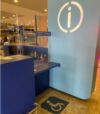Ponto de atendimento com sinalização tátil no piso para pessoas com deficiência, com balcão rebaixado e comunicação visual com o símbolo de informação em destaque na parede azul.