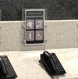 Área com telefones de atendimento sobre o balcão e, ao fundo, uma placa com quatro ícones, incluindo o símbolo do aro magnético (orelha com linha e “T”), indicando que aquele setor oferece assistência acessível a diferentes deficiências.