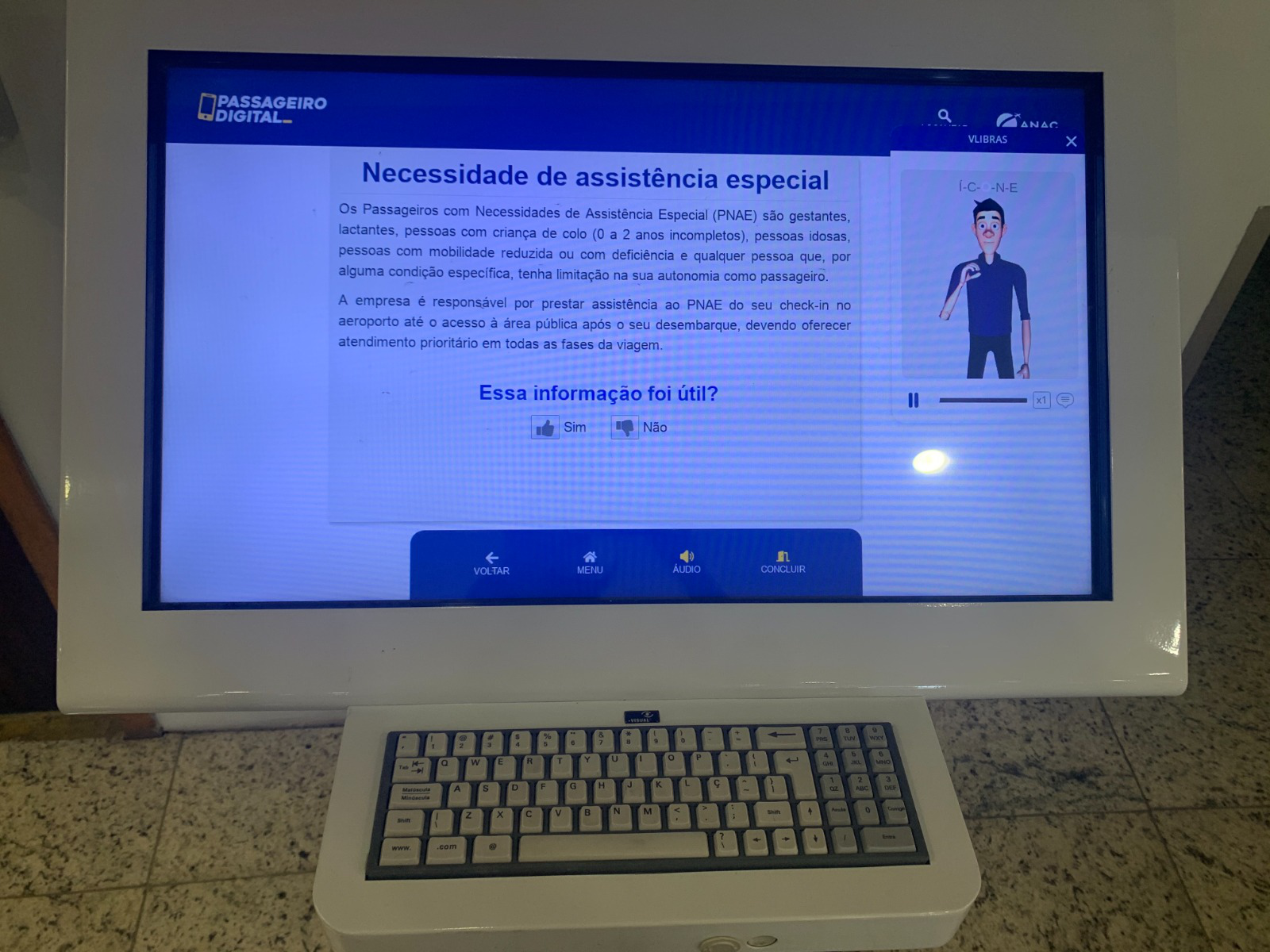 Totem informativo digital com teclado físico e tela exibindo informações sobre “Necessidade de assistência especial”, acompanhadas de uma janela de Libras no canto direito, com um intérprete virtual sinalizando o conteúdo.