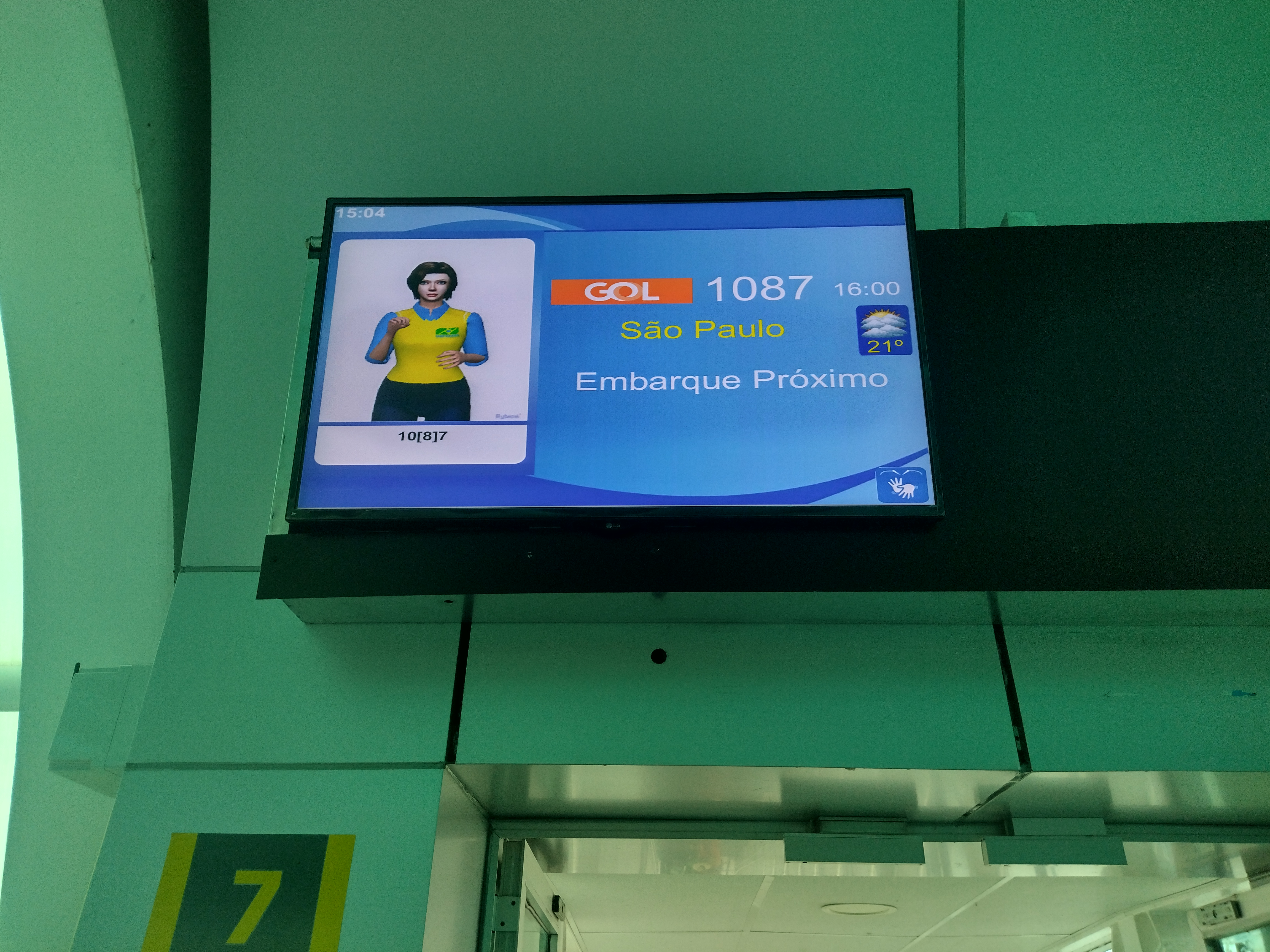 Tela suspensa exibindo dados de voo (voo GOL 1087 para São Paulo) com uma janela de Libras do lado esquerdo da tela, onde uma intérprete sinaliza a mensagem “Embarque Próximo”.