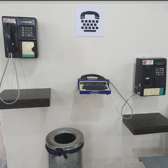Vista frontal de um conjunto de telefones públicos fixados na parede, com telefone convencional à esquerda, telefone TDD (para comunicação com pessoas surdas) ao centro e outro convencional à direita, todos em prateleiras acessíveis.