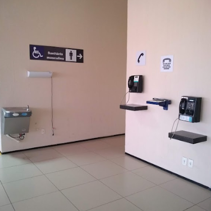 Ambiente com bebedouro acessível, placa de sanitário masculino com símbolo de acessibilidade e conjunto de telefones acessíveis fixados na parede em prateleiras a alturas adequadas, incluindo um TDD no centro.