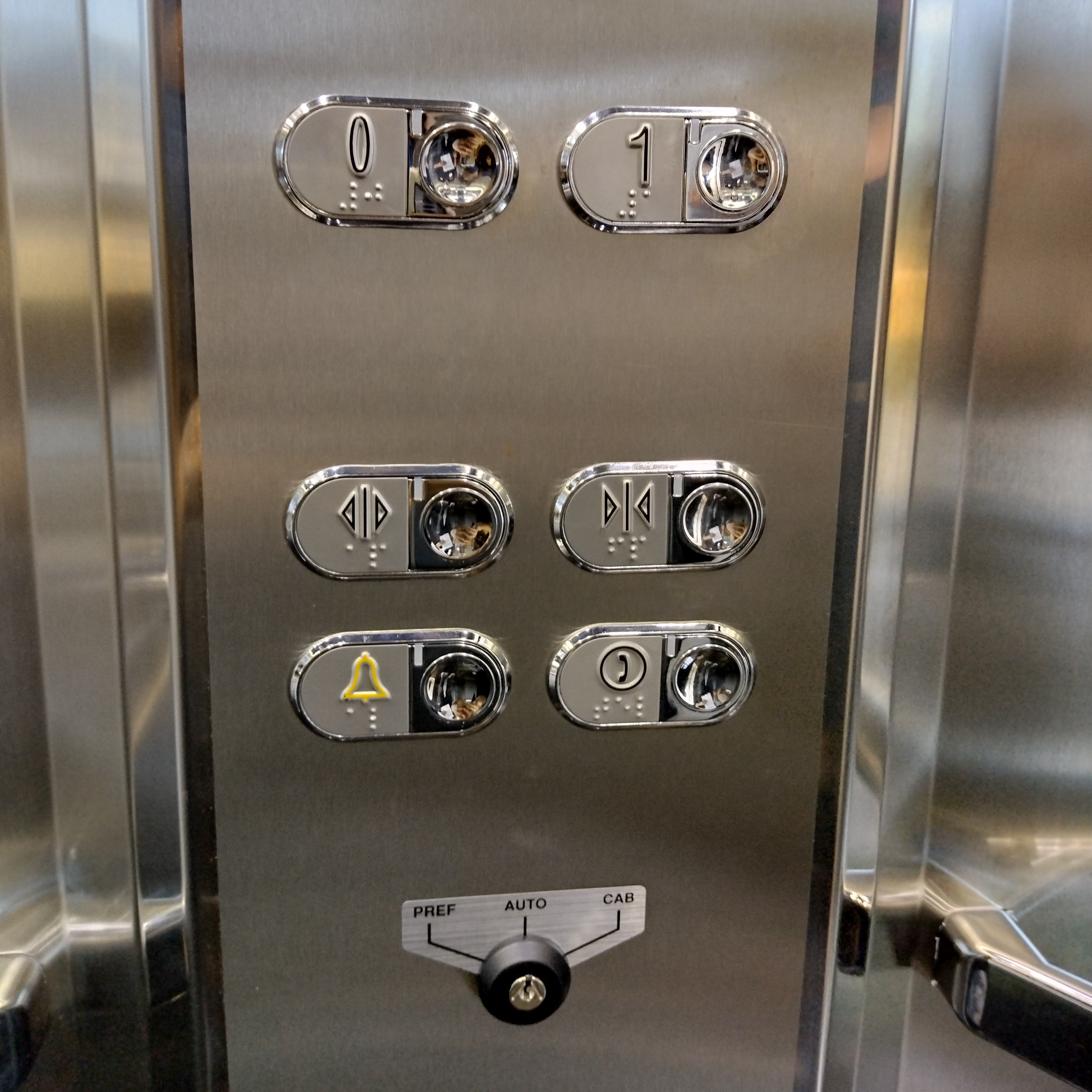 Painel de elevador com botões em alto-relevo e em braille, com indicação de andares, abrir/fechar portas e alarme. O botão de alarme tem iluminação amarela.