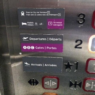Painel de elevador com botões visuais e táteis em braille. Cada botão tem numeração de andares e setas de direção, com informações em inglês e francês.