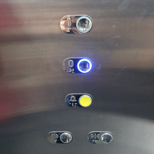 Painel de elevador com botões acessíveis, incluindo iluminação de chamada, botão de alarme amarelo, e etiquetas em braille em cada botão.