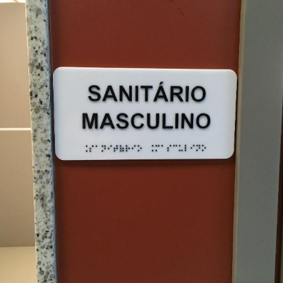 Placa fixada na parede ao lado de uma porta, com a inscrição “Sanitário Masculino” em letras ampliadas e Braille logo abaixo.