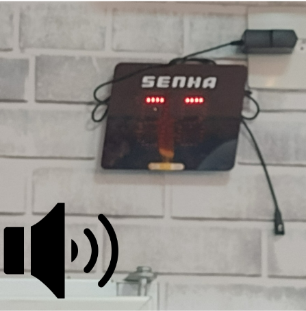 Painel eletrônico de senhas fixado na parede, com números iluminados em LED e ícone de alto-falante na imagem, indicando que o sistema possui chamada sonora para garantir acessibilidade no atendimento.