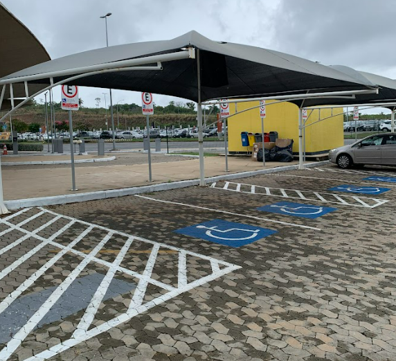 Vagas externas acessíveis sob cobertura, com piso em blocos intertravados e demarcações brancas e azuis indicando reserva para pessoas com deficiência.