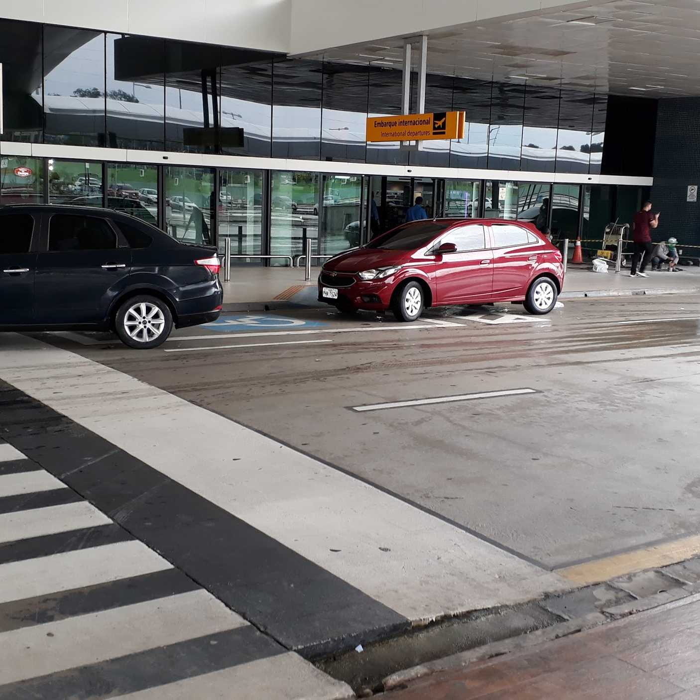 Área externa de embarque de um aeroporto com rebaixamento de calçada, piso tátil direcional e vaga reservada para pessoas com deficiência em frente à entrada principal.