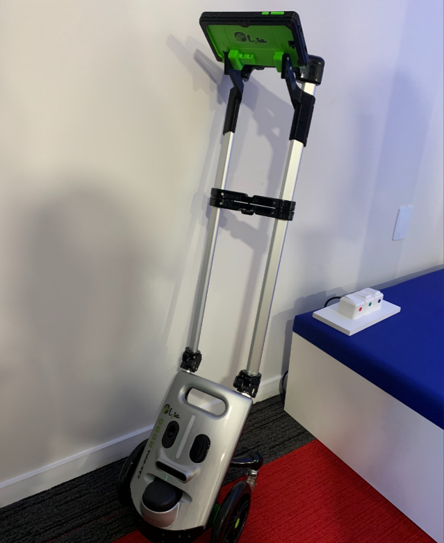 Equipamento vertical com rodas, estrutura metálica e tela na parte superior, apoiado junto à parede. Trata-se de um dispositivo eletrônico assistivo que pode acompanhar o passageiro e fornecer orientação autônoma pelo ambiente aeroportuário.