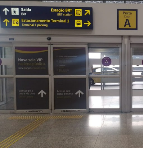 Porta de vidro automática no aeroporto, com piso tátil direcional amarelo à frente e sinalizações indicativas de setores como estacionamento, estação BRT e saída, localizados acima da porta.