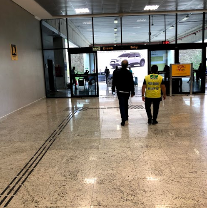Entrada ampla com portas de vidro automáticas e piso tátil direcional no saguão do aeroporto. Duas pessoas caminham em direção à saída, onde há sinalização visível com setas e informações.