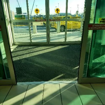 Vista interna de uma porta automática transparente no aeroporto, com acesso nivelado ao meio-fio externo, onde se vê um táxi amarelo aguardando. Há sinalização de direção visível na parte superior das portas.