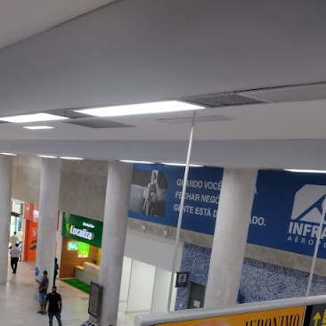 Vista ampla de saguão de aeroporto com pé direito alto e iluminação artificial no teto. O ambiente conta com áreas de atendimento ao público e locadoras de veículos. A iluminação é uniforme, garantindo boa visibilidade em toda a área.