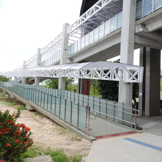 Corredor externo coberto, com rampa larga protegida por guarda-corpo de vidro e aço inox. A estrutura está conectada ao edifício superior, com percurso contínuo, seguro e sem obstruções. O piso é plano e a inclinação da rampa segue os padrões normativos.