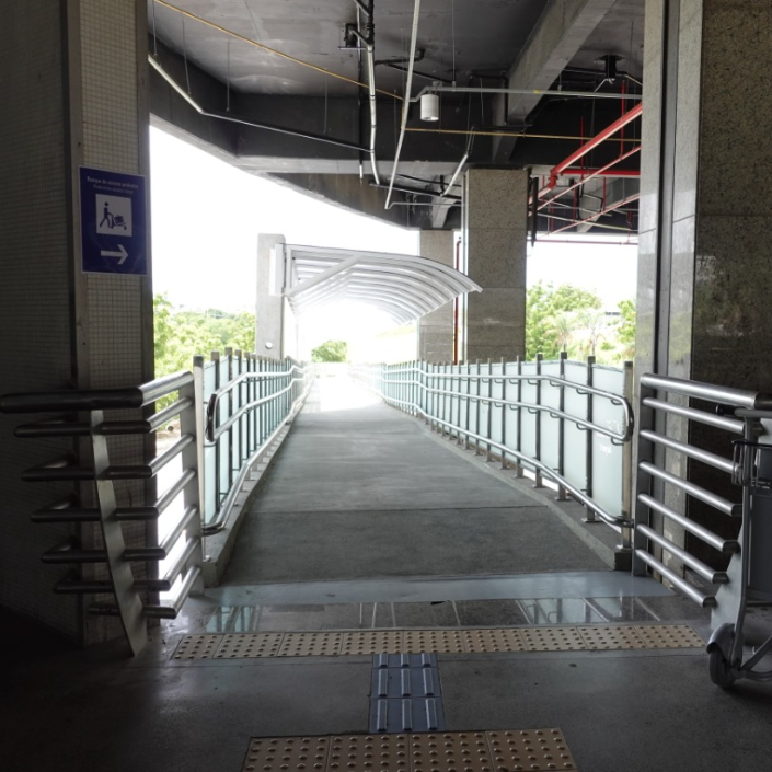 Vista interna de um corredor acessível em nível elevado, com piso regular e contínuo. O espaço possui corrimãos bilaterais em duas alturas, além de sinalização tátil direcional na entrada e saída, garantindo a acessibilidade para pessoas com deficiência.