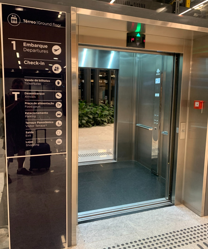 Vista externa de um elevador acessível em funcionamento, com a porta aberta no piso térreo de um aeroporto. Ao lado, há uma placa vertical com indicação clara dos andares e serviços disponíveis, com pictogramas acessíveis e informações bilíngues (português e inglês). Há também piso tátil direcional.