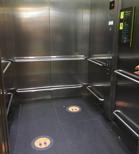 Interior de um elevador amplo, com barras de apoio em todas as paredes, incluindo ao lado do painel de botões. O espaço permite a circulação e manobra de cadeira de rodas. Adesivos no chão indicam marcação para distanciamento físico.