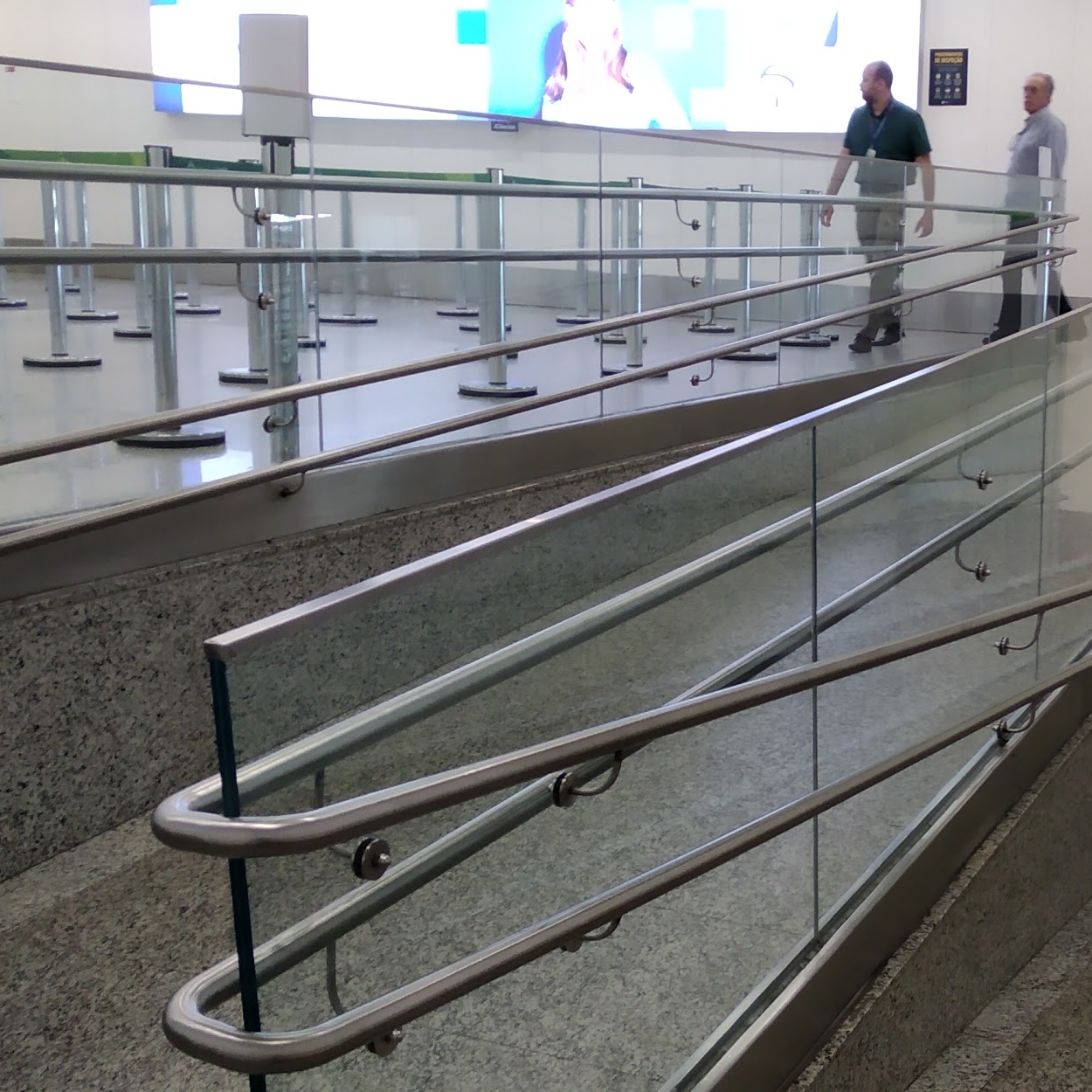 Rampa acessível com piso antiderrapante e guarda-corpo de vidro, localizada em área interna de aeroporto, com corrimãos duplos em ambas as laterais, em diferentes alturas, conforme norma de acessibilidade.