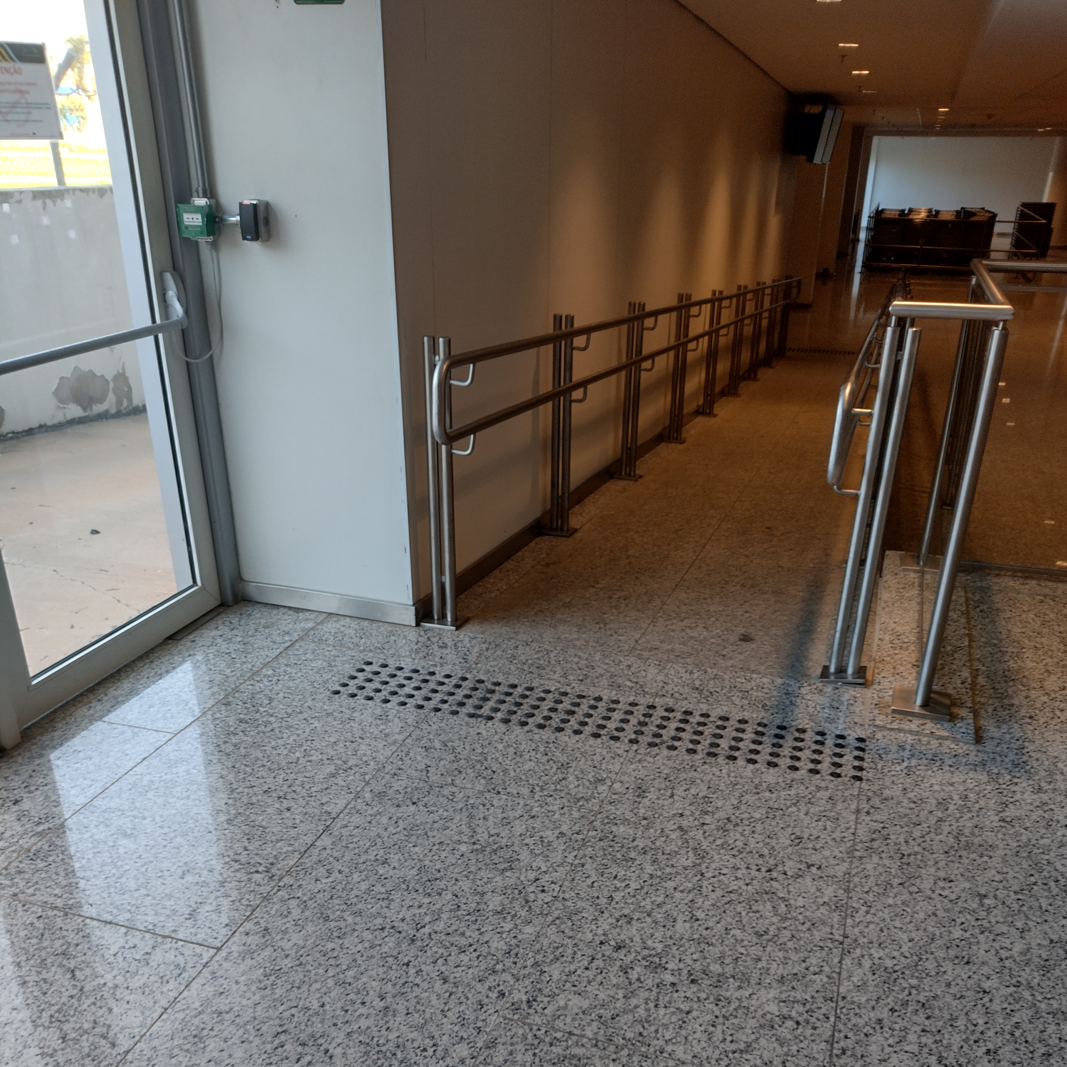 Rampa de acesso próxima a uma saída lateral do aeroporto, com piso tátil de alerta na entrada e corrimãos duplos contínuos em aço inox dos dois lados, atendendo às exigências de acessibilidade normativa.