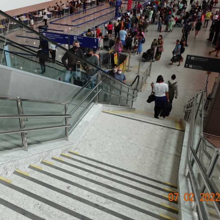 Escadaria ampla em terminal de aeroporto, com corrimãos em dois níveis, guarda-corpos laterais, piso tátil de alerta no início da escada e sinalização visual de contraste nos degraus.