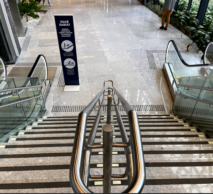 Escadaria moderna com piso em granito, corrimãos duplos e faixa de sinalização de segurança nos degraus. Ao lado, escadas rolantes e vegetação decorativa interna.